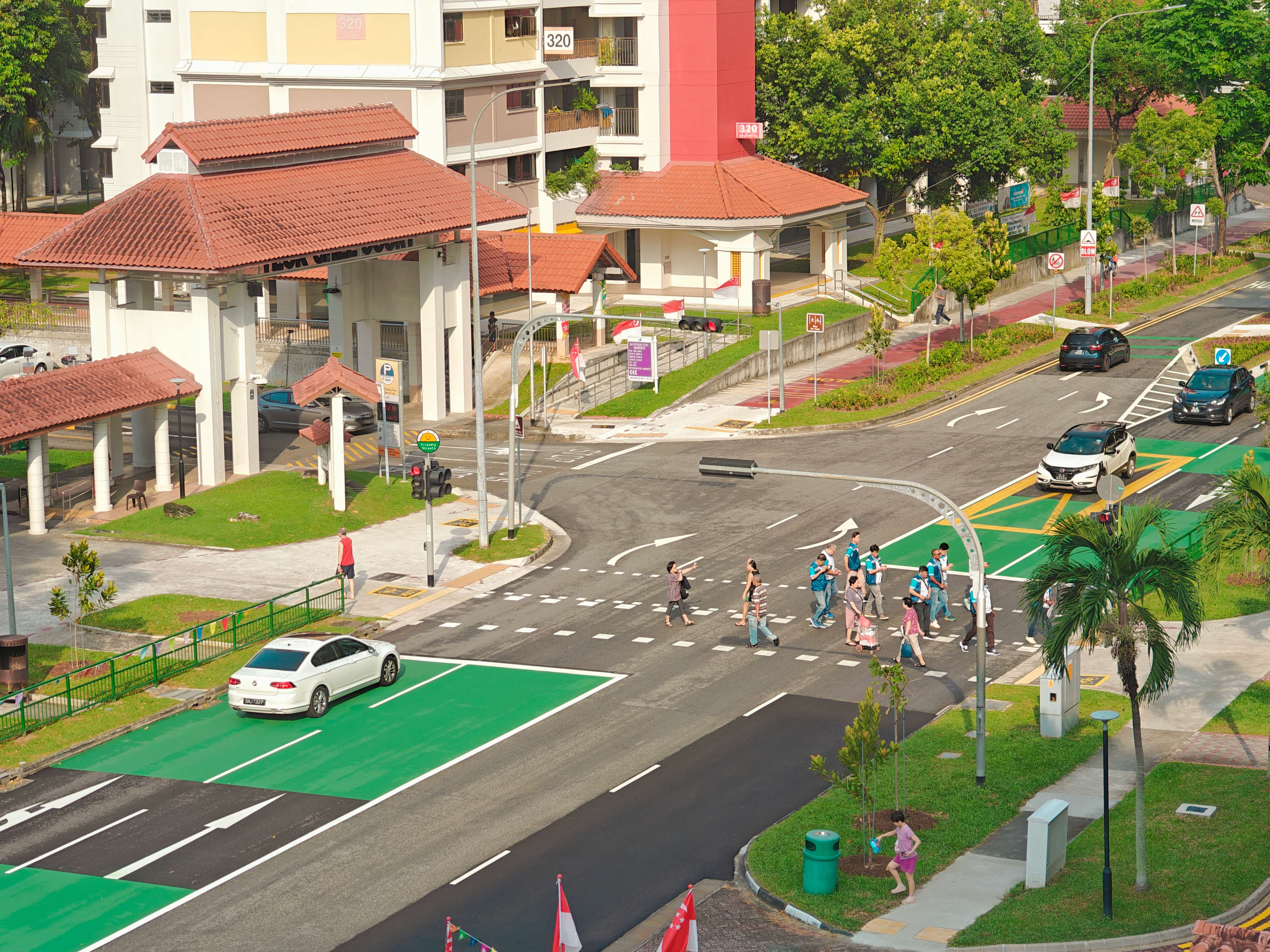 An aerial view shows a group of people in a pedestrian crossing on a road surrounded by buildings and greenery in a Singaporean neighborhood.